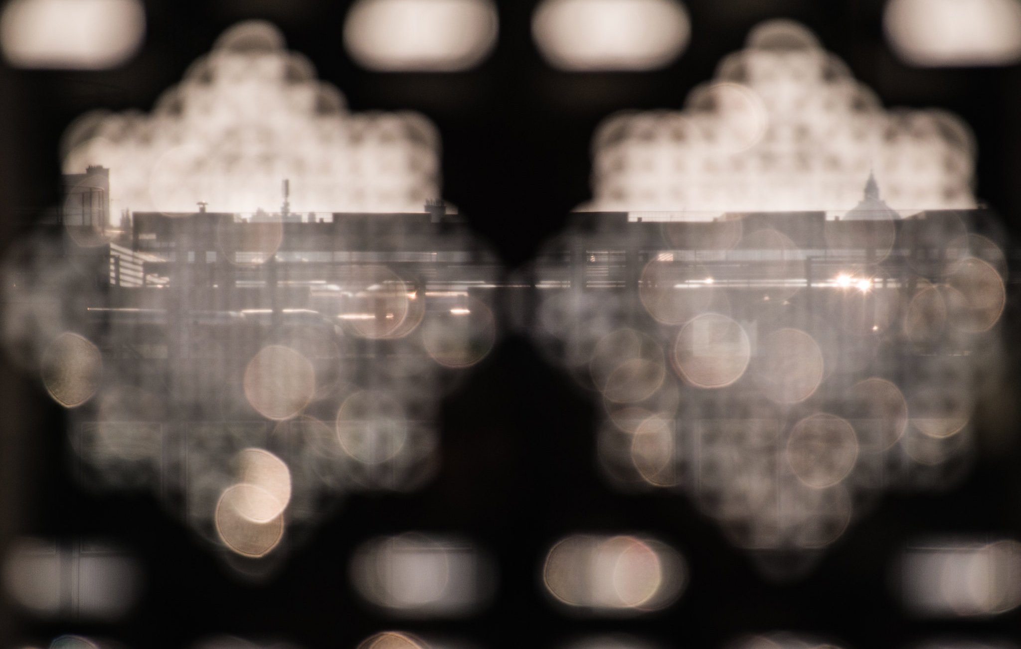 L'image montre une vue floue à travers une structure découpée, de formes géométriques. On peut apercevoir des silhouettes de bâtiments en arrière-plan, avec une lumière réfléchie qui crée des éclats. L'atmosphère générale semble douce et légèrement mystique, accentuée par les jeux de lumière et de flou. Les détails précis des éléments au loin ne sont pas nettement visibles, ce qui ajoute à l'effet artistique de la composition.