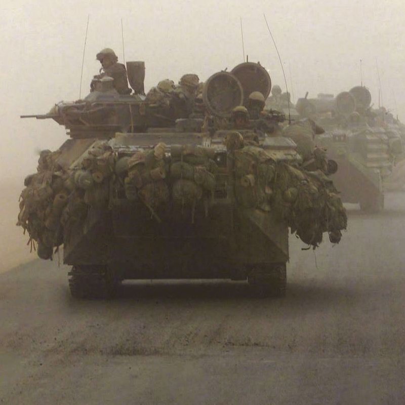 The image depicts military vehicles, specifically tanks, moving through a dusty and foggy environment. The tanks are heavily laden with gear and equipment, indicating they are prepared for deployment or combat. The visibility in the scene is limited due to the dust, which creates a muted atmosphere. Soldiers can be seen perched on top of the tanks, suggesting readiness and vigilance as they navigate the terrain. The overall mood conveys a sense of military operation amid challenging conditions.