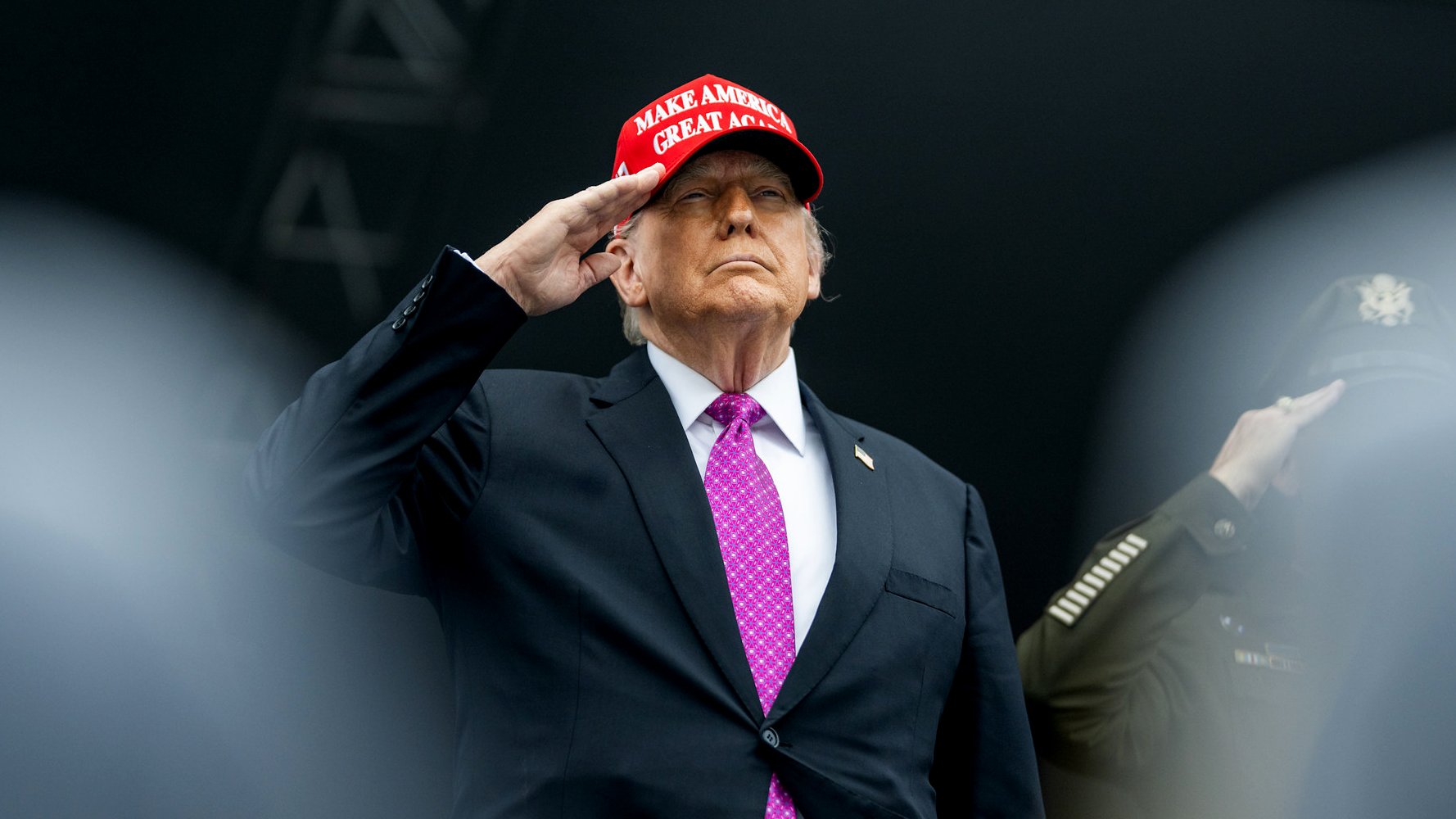 مردی با کلاهی قرمز و پیراهن مشکی، با دست بر پیشانی ایستاده و سلام می‌کند.