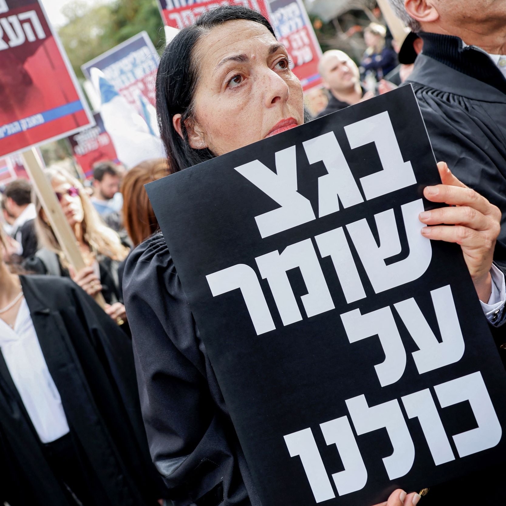 در تصویر، گروهی از افراد در حال برگزاری یک تظاهرات هستند. زنان و مردان با لباس‌های تیره و جعبه‌های دست‌نوشته‌ای در دست دارند که بر روی آنها نوشته‌هایی به زبان عبری دیده می‌شود. یکی از زنان در جلو تصویر، به وضوح در حال نگاه کردن به دوربین است و یک پلاکارد بزرگ با نوشته‌ای مشکی در دست دارد. فضای کلی تظاهرات نشان‌دهنده همبستگی و اعتراض مردم به یک موضوع خاص است.