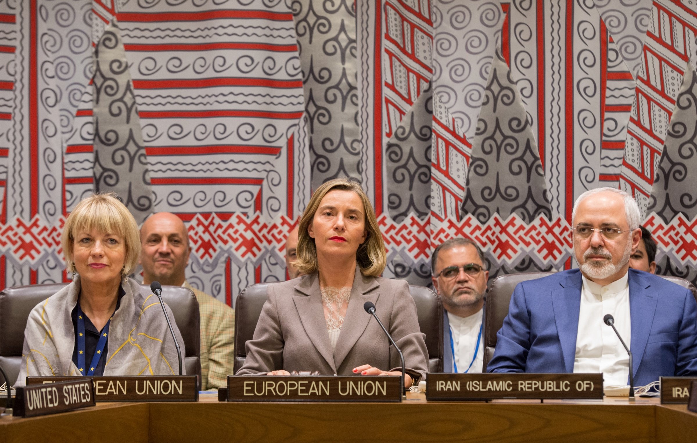 L'image montre une réunion internationale avec plusieurs personnes assises à une table. On peut voir des panneaux identifiant les différents participants, tels que l'Union européenne et l'Iran. Trois figures centrales semblent être des représentants importants, avec des expressions sérieuses. En arrière-plan, il y a des individus qui écoutent attentivement. Le décor est riche et coloré, avec des motifs géométriques sur le mur derrière eux, suggérant un cadre formel pour des discussions diplomatiques.