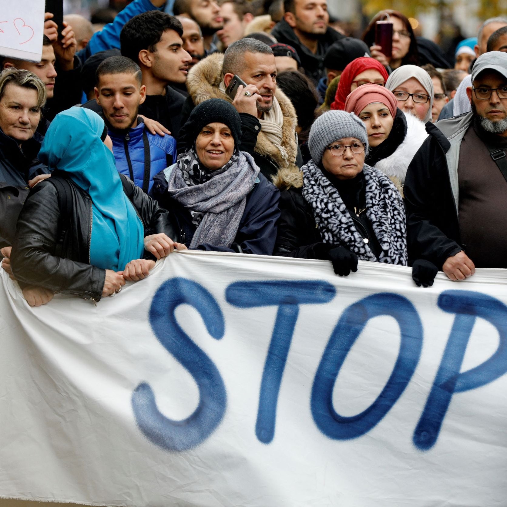 L'image montre une foule de personnes rassemblées pour une manifestation. Elles tiennent des pancartes, dont l'une affiche le mot "STOP" écrit en grandes lettres. Les participants portent des vêtements variés, certains ayant des écharpes ou des chapeaux, ce qui suggère qu'il pourrait faire frais. L'atmosphère semble engagée, avec des visages sérieux ou déterminés, reflétant un fort sentiment de solidarité pour la cause qu'ils soutiennent.