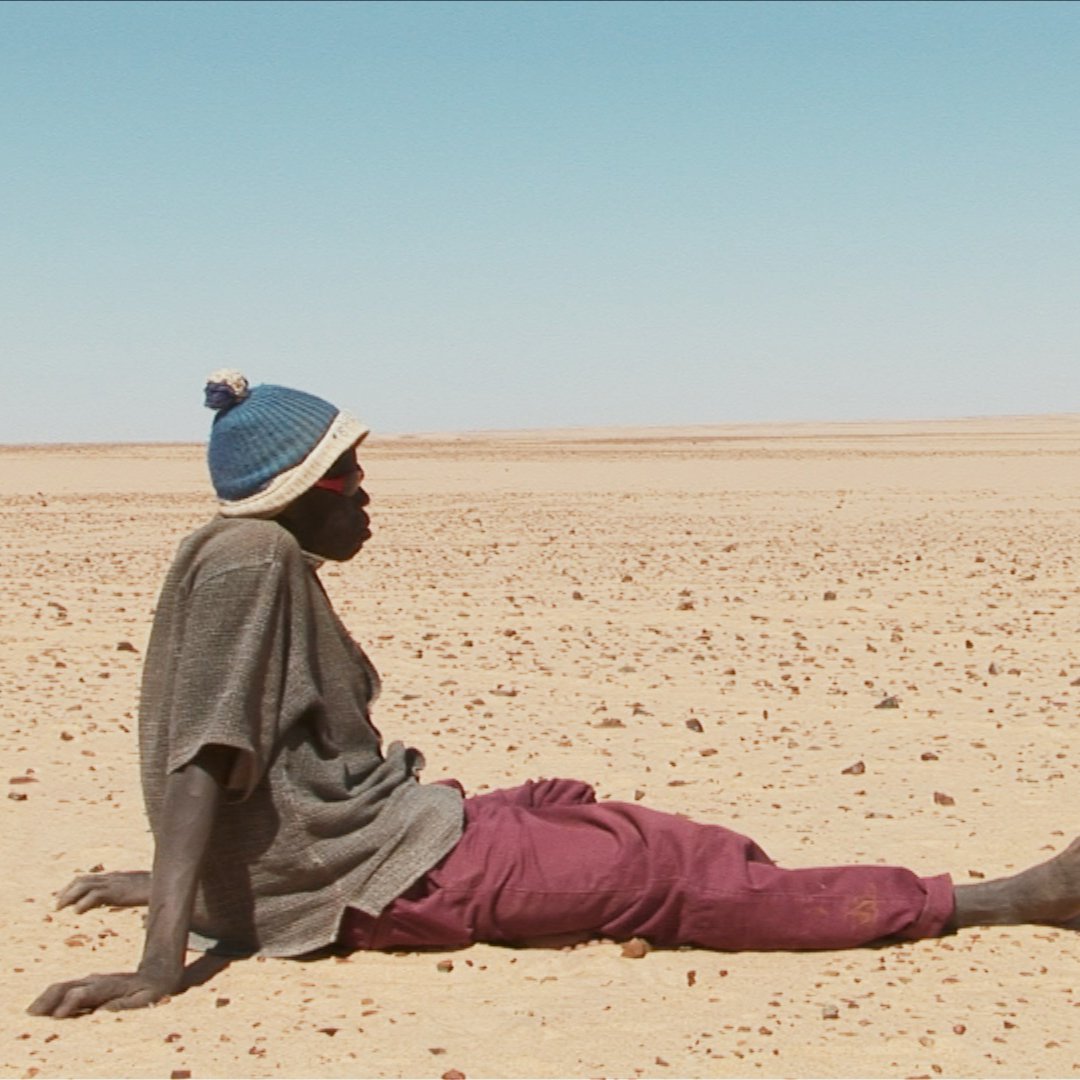 L'image montre un homme assis sur le sol désertique. Il porte un pull avec un bonnet et un pantalon de couleur prune. Le paysage autour de lui est vaste et aride, constitué principalement de sable et de petites pierres, s'étendant à perte de vue sous un ciel clair. L'atmosphère semble à la fois isolée et silencieuse, évoquant une sensation de solitude dans cet environnement désertique.