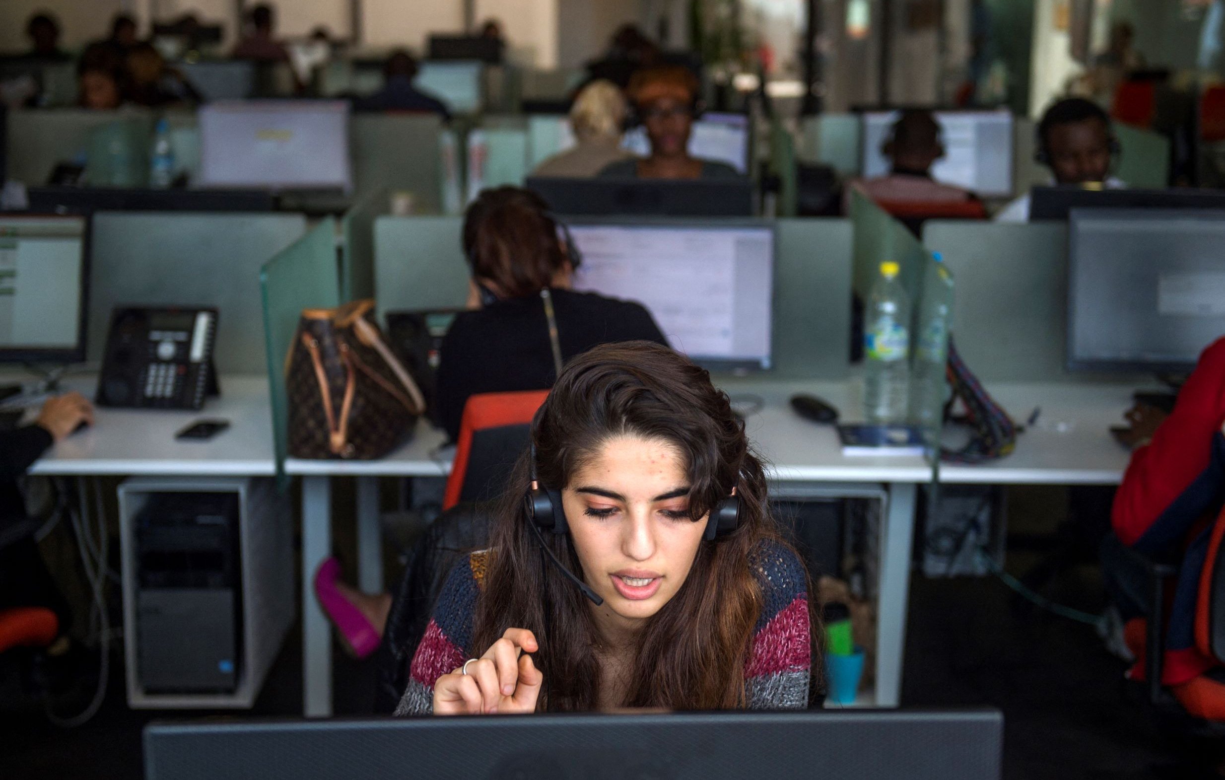 تظهر الصورة مكتبا حديثا في بيئة عمل، حيث توجد مجموعة من الأشخاص يعملون في مراكز اتصال. في المقدمة، تجلس شابة ترتدي سماعة رأس وتتحدث إلى ميكروفون، بينما تركز على شاشة الكمبيوتر أمامها. خلفها، يمكن رؤية زملاء آخرين يعملون، كلٌ منهم متواجد أمام حاسوبه. يظهر في الخلفية بعض الأغراض الشخصية مثل الحقائب، مما يضيف طابعاً إنسانياً للمكان. الإضاءة في المكتب طبيعية وتساهم في خلق جو عملي.