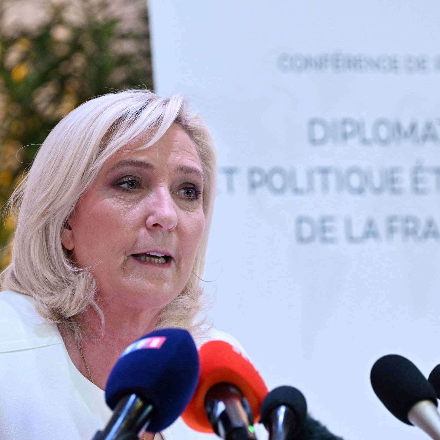 L'image montre une femme qui s'exprime lors d'une conférence de presse. Elle a des cheveux blonds et porte une veste claire. Au fond, on peut voir un panneau qui indique "CONFERENCE DE PRESSE" ainsi que le titre "DIPLOMATIE ET POLITIQUE ÉTRANGÈRE DE LA FRANCE". Plusieurs microphones sont disposés devant elle, suggérant qu'elle s'adresse aux médias. L'ambiance semble formelle et professionnelle.