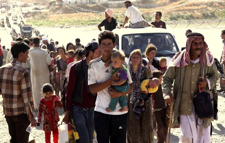 L'image montre un grand groupe de personnes se déplaçant, probablement dans un contexte de crise. On voit des adultes et des enfants marchant, avec des expressions sérieuses sur leurs visages. Certains portent des objets, et il y a une camionnette dans le fond. L'environnement semble désolé, avec peu d'infrastructures visibles. Cette scène évoque des thèmes de déplacement et de lutte, typiques de situations de conflit ou de catastrophe.