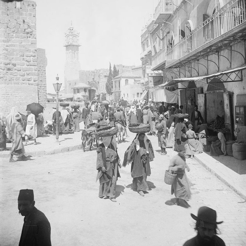 L'image montre une scène animée à la porte de Jaffa à Jérusalem. On y voit de nombreuses personnes en mouvement, portant des vêtements traditionnels. Certains portent des charges sur leur tête, tandis que d'autres se déplacent dans la rue. Des bâtiments historiques se dressent en arrière-plan, et l'atmosphère semble occuper un marché ou un lieu de rencontre. La lumière est vive, ce qui donne une impression de chaleur et d'activité.