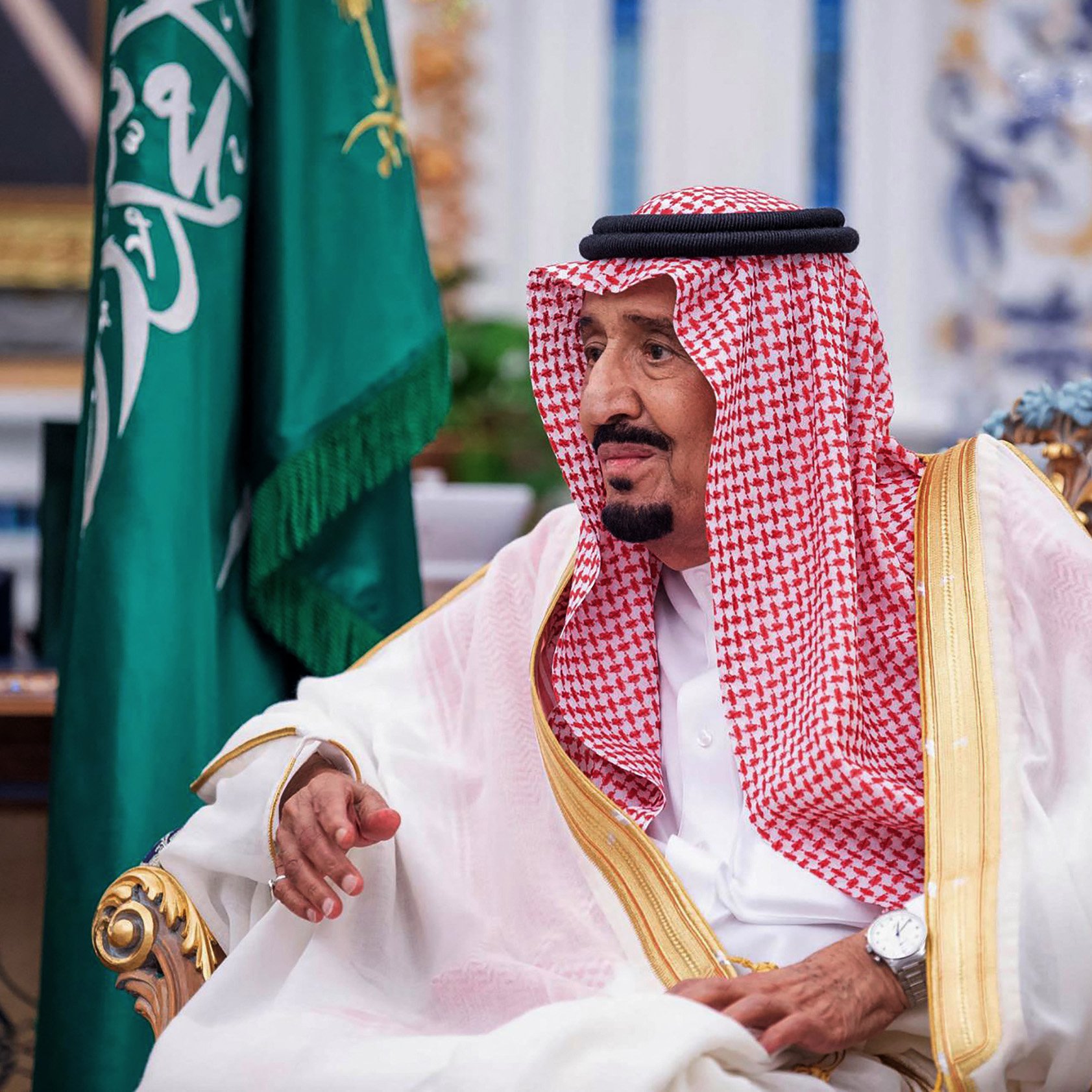 تظهر في الصورة شخصية ترتدي ملابس تقليدية سعودية، تتضمن الإزار والعباءة، كما أن هناك شالًا على رأسه. يجلس على كرسي فخم في بيئة ذات تفاصيل تراثية. خلفه توجد علم المملكة العربية السعودية، مما يدل على السياق الرسمي والمناسبات المهمة. تعكس تعابير وجهه تركيزًا واهتمامًا، كما يظهر الاهتمام بالتصميم والزخارف المحيطة به.