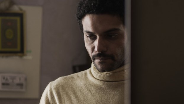 L'image montre un homme avec des cheveux bouclés et une moustache, portant un pull à col roulé de couleur claire. Il semble pensif, regardant vers le bas, avec une expression de réflexion ou de sérieux. En arrière-plan, on aperçoit des éléments de décoration intérieure, mais les détails sont flous. L'atmosphère de l'image dégage une certaine intimité ou mélancolie.