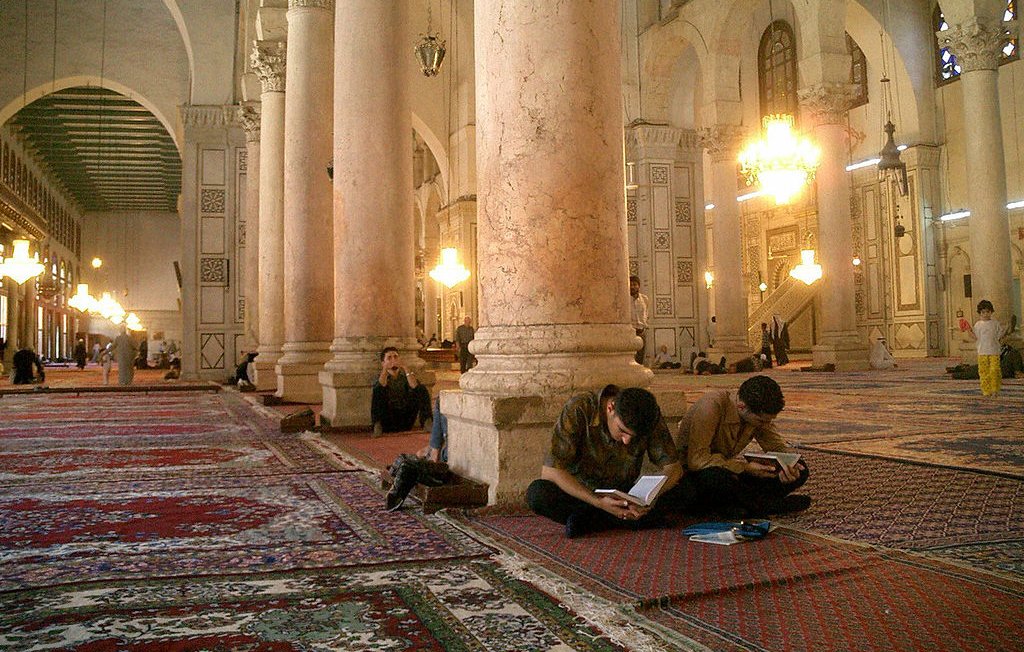 L'image montre l'intérieur d'une grande mosquée, caractérisée par de hauts plafonds et de majestueuses colonnes. Le sol est recouvert de tapis, et plusieurs personnes sont assises sur le sol, certaines concentrées sur leurs ordinateurs portables, tandis que d'autres semblent prier ou méditer. La lumière naturelle entre par les fenêtres, créant une ambiance calme et sereine. Des lustres pendent du plafond, ajoutant une touche d'élégance à cet espace sacré.
