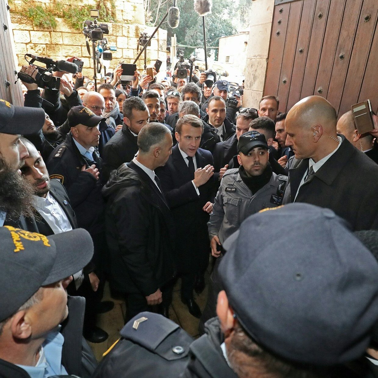 تصویر گروهی از افراد را نشان می‌دهد که در یک محیط شلوغ با امنیت زیاد در حال گفتگو هستند. تعدادی از افراد یونیفورم‌پوش به نظر می‌رسد که نگهبان هستند و در حال محافظت از یک فرد دیگر هستند. افراد دیگر در اطراف آن‌ها ایستاده‌اند و به نظر می‌رسد که برخی از آن‌ها مشغول ضبط یا عکاسی از این صحنه هستند. فضا به شدت پر ازدحام و حاکی از توجه رسانه‌ای است.