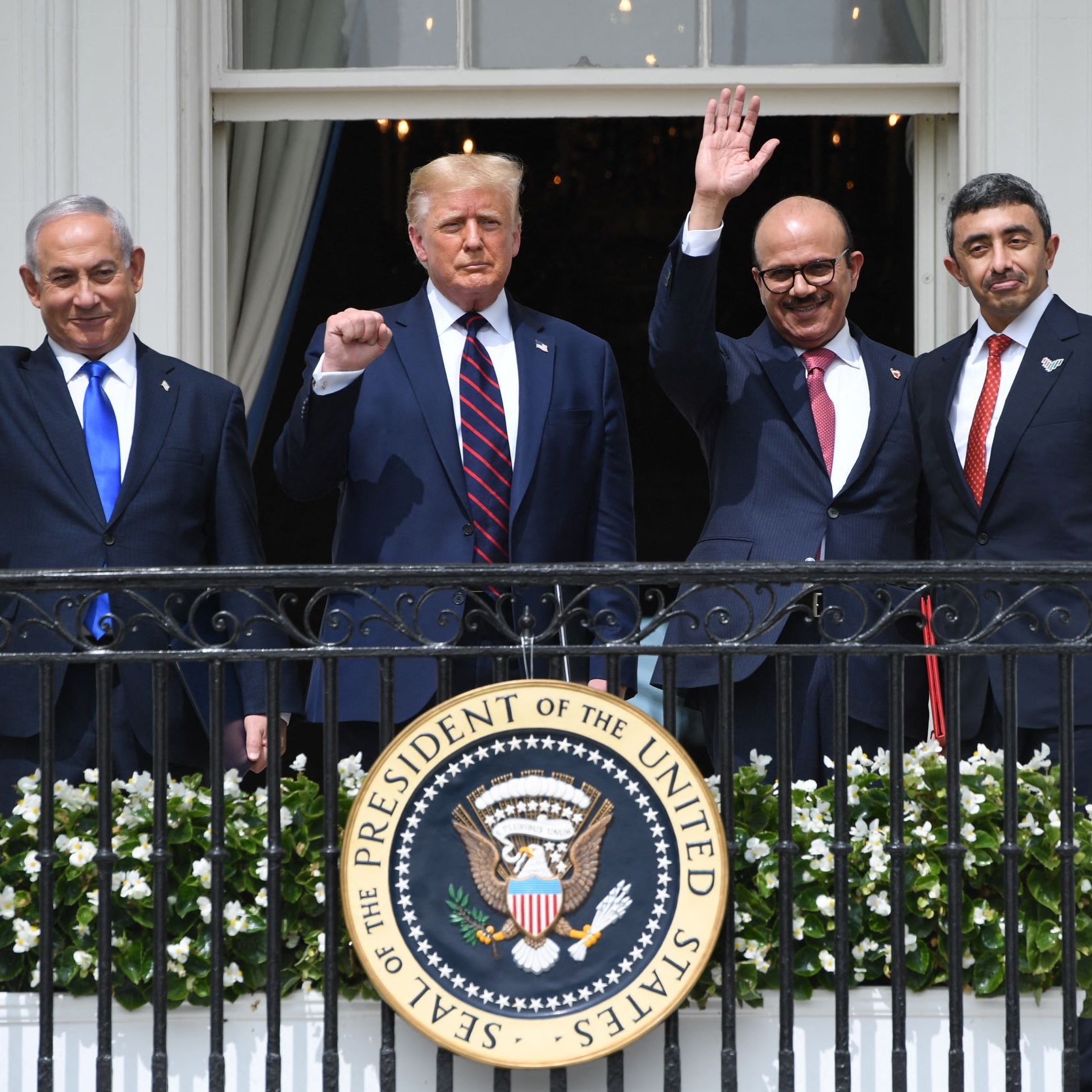 L'image montre quatre hommes debout sur un balcon de la Maison Blanche, levant la main en signe de salutation. Ils semblent être en train de célébrer un moment important, probablement lié à des événements diplomatiques. L'un des hommes porte un costume sombre avec une cravate rouge, tandis que les autres affichent également des tenues formelles. En arrière-plan, on peut voir un environnement architectural caractéristique de la Maison Blanche, et un grand sceau du Président des États-Unis est en évidence sur la balustrade.