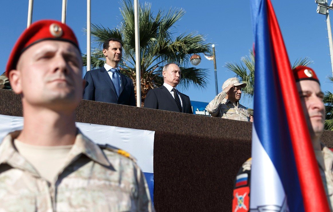 L'image montre un groupe de personnes, dont deux figures de haut rang, sur une plateforme en extérieur, avec un fond de palmiers et un ciel bleu. Au premier plan, des soldats en uniforme, probablement d'une unité militaire, sont présents, portant des drapeaux. Les expressions des personnes sur la plateforme semblent sérieuses et concentrées, indiquant un événement officiel ou militaire.