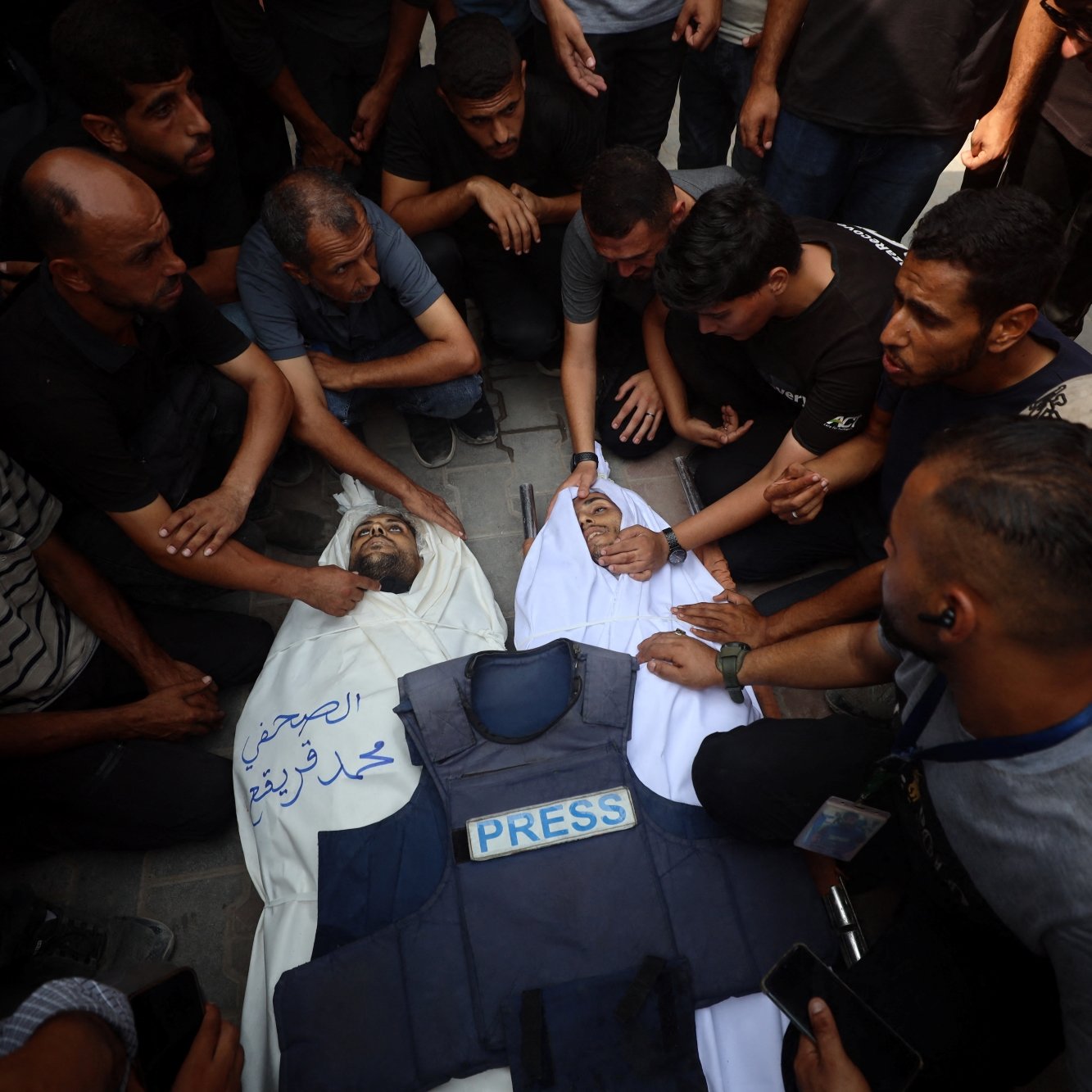 Des hommes entourent un corps recouvert, en deuil, avec une veste de presse à côté.