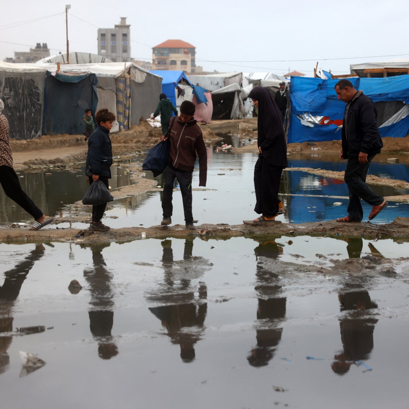 در تصویر، گروهی از مردم در یک منطقه نامناسب عبور می‌کنند. زیر پاهای آن‌ها آب جمع شده و انعکاس آن‌ها در آب دیده می‌شود. چادرهای موقتی در زمینه وجود دارند که به وضوح مشخص است. آسمان ابری و فضای کلی تصویر حاکی از شرایط سخت و نامساعد است. افراد در حال حرکت به سمت مقصدی مشخص هستند و لباس‌های آن‌ها نشان‌دهنده وضعیت اقتصادی و اجتماعی‌اشان است.