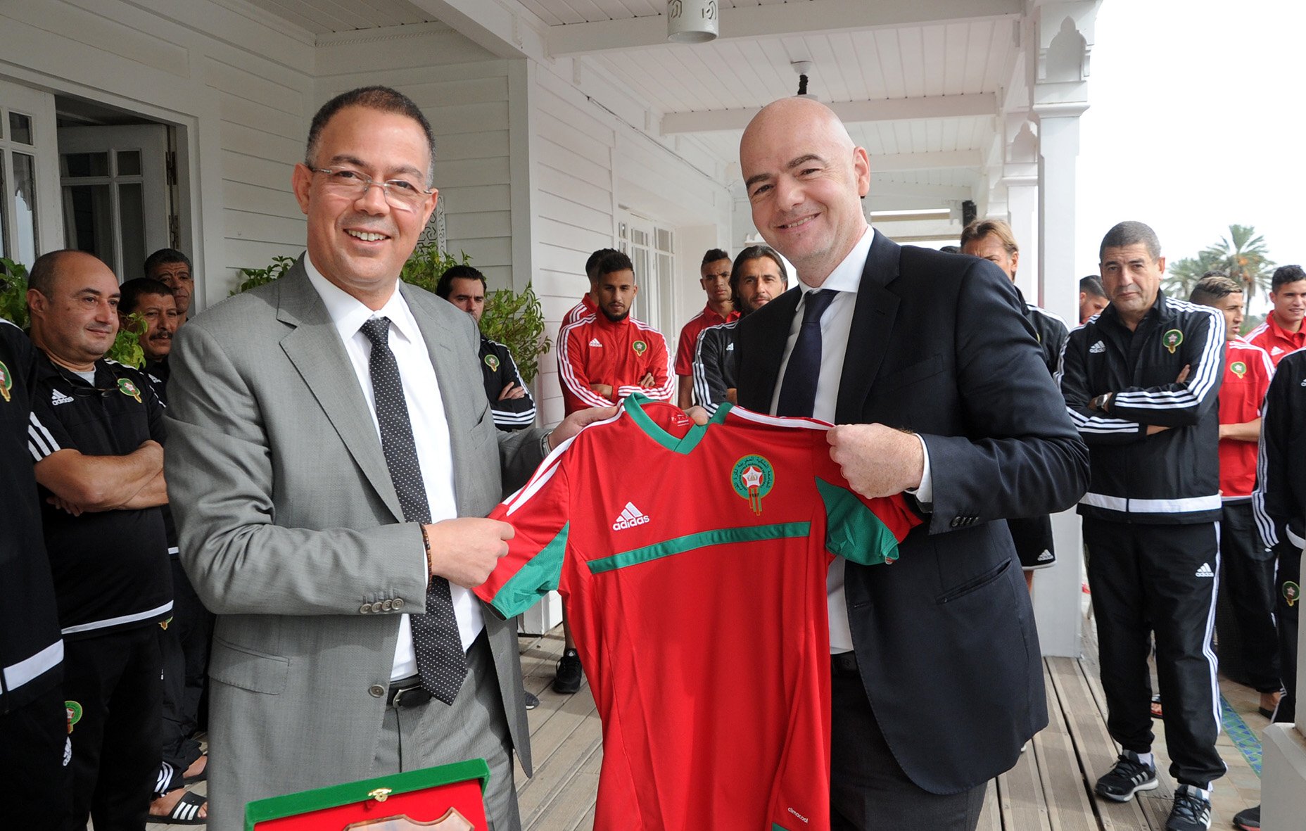 L'image montre deux hommes souriants qui se tiennent devant un groupe de personnes en arrière-plan. L'un d'eux porte un costume gris et tient un maillot de football rouge et vert, probablement aux couleurs de l'équipe nationale marocaine. L'autre homme, en costume sombre, semble également heureux et participe à la remise de ce maillot. Les autres personnes en arrière-plan portent des ensembles sportifs noirs avec des détails blancs. L'ambiance suggère un moment de célébration ou d'échange, probablement lié au football.