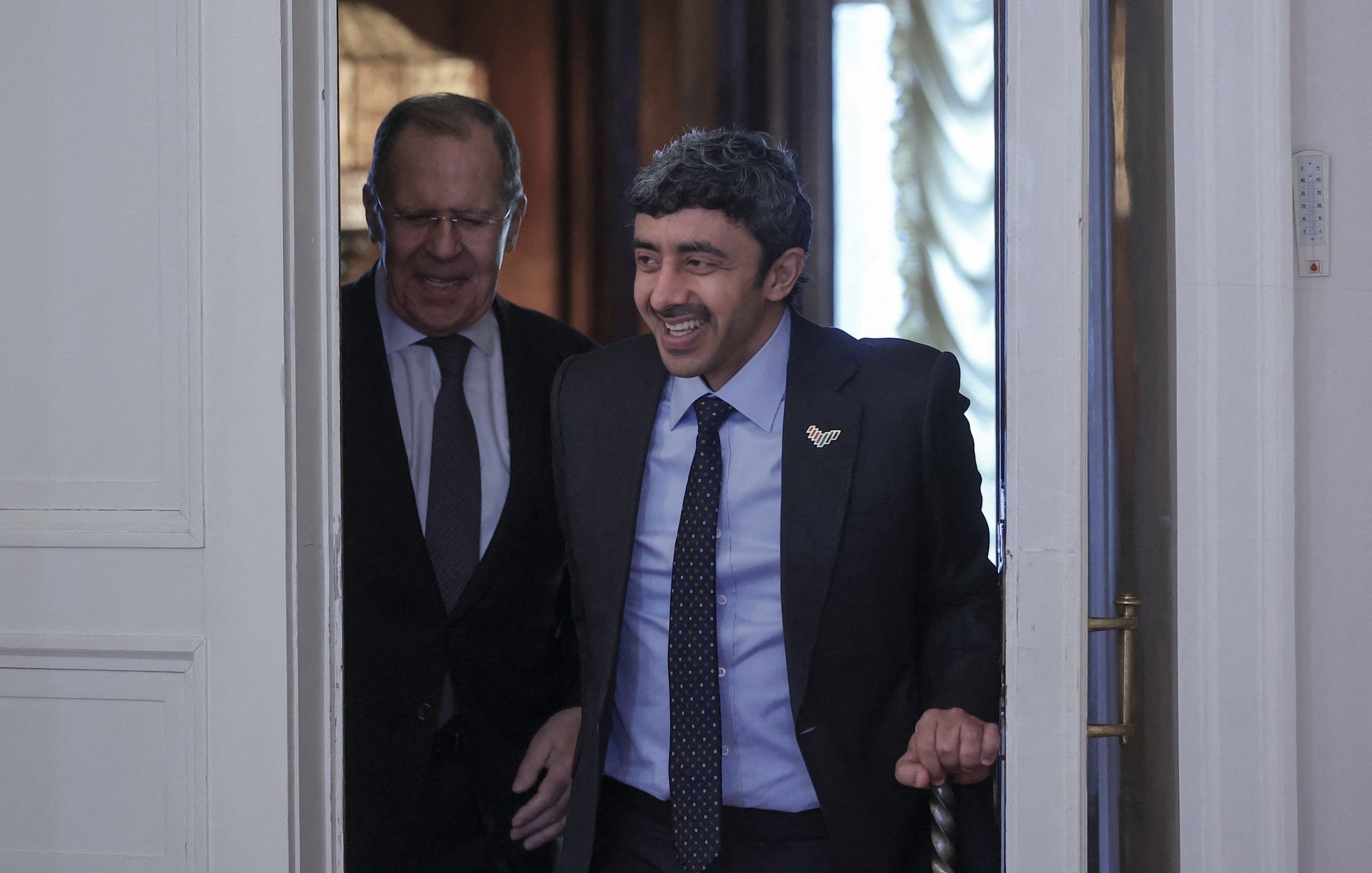 The image shows two men entering a room together. One man is smiling and appears to be in a suit, indicating a formal occasion. The other man, who seems more serious, is walking closely behind. The setting looks elegant, likely a governmental or official location, with decorative elements visible in the background. The overall mood suggests a friendly or cordial interaction between the two individuals.