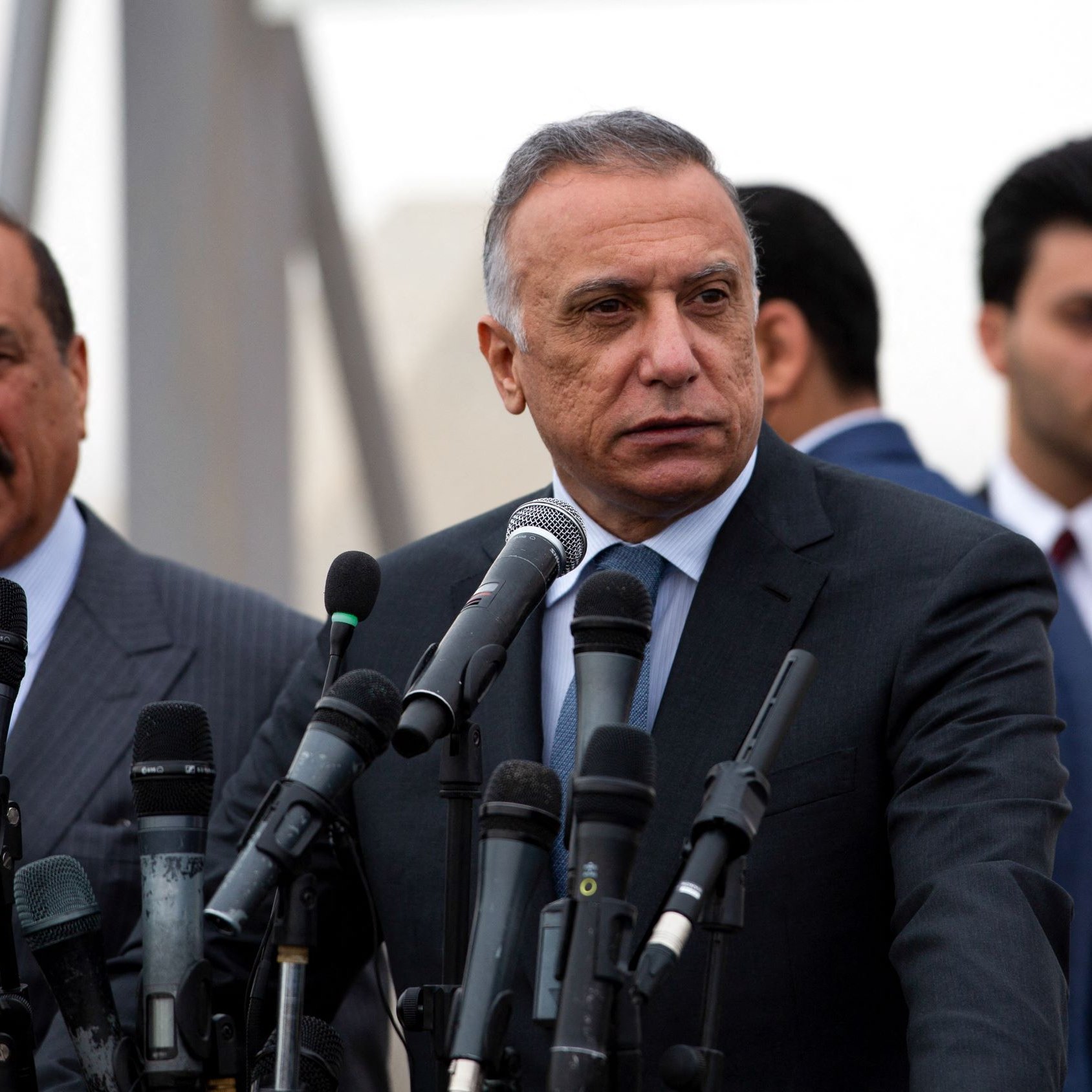 L'image montre un groupe d'hommes lors d'une conférence de presse. Au premier plan, deux hommes sont debout devant plusieurs microphones. L'un d'eux, qui se tient à gauche, porte un costume rayé et semble s'adresser à l'audience. L'autre homme, au centre, est également en costume et adopte une pose sérieuse, regardant au loin. En arrière-plan, d'autres hommes en costumes sont visibles, ajoutant à l'ambiance formelle de l'événement. L'environnement semble légèrement brumeux.