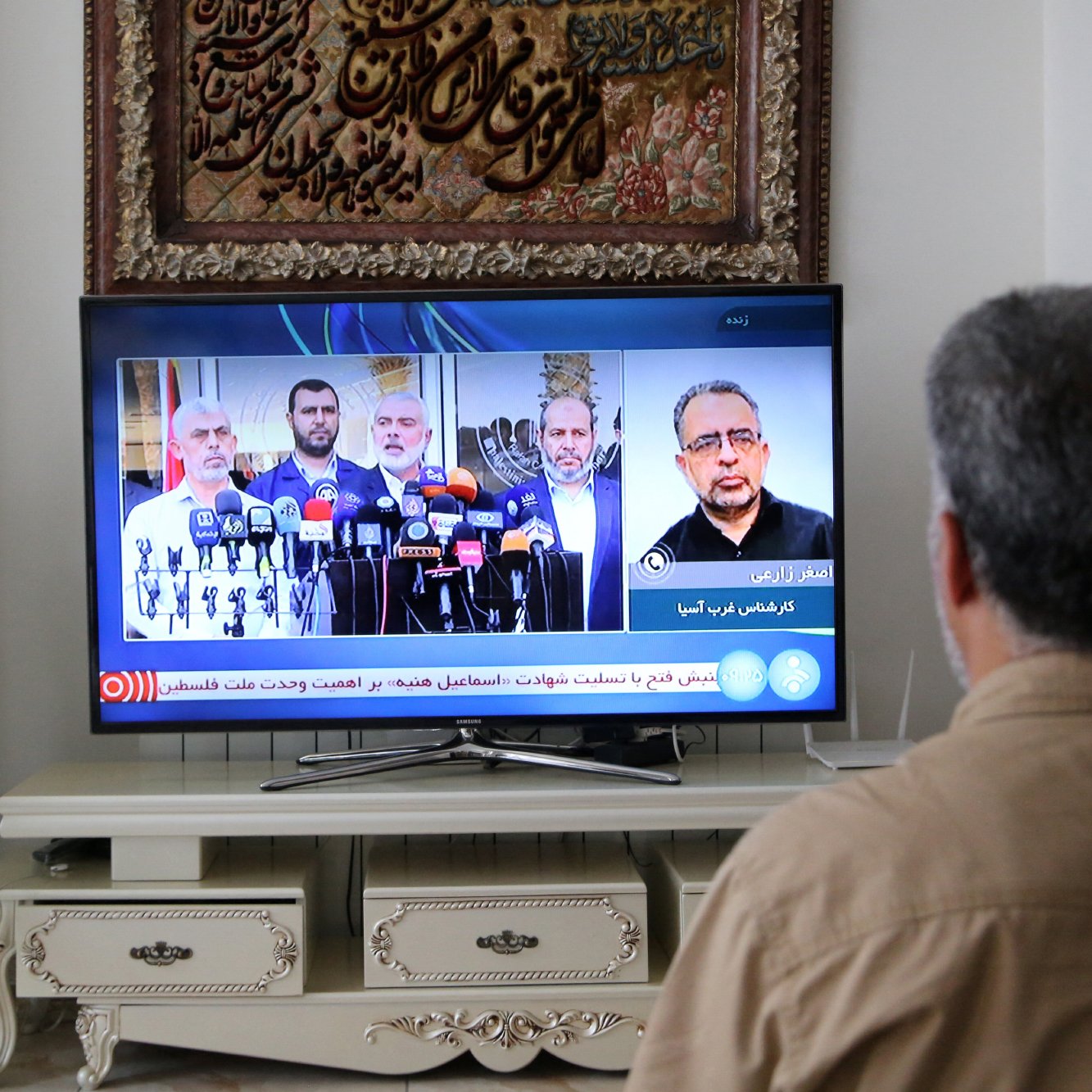 L'image montre une personne assise devant un téléviseur, en train de regarder une retransmission en direct. Sur l'écran, on peut voir un groupe de personnes qui semblent donner une conférence de presse, entourées de microphones. Le décor derrière inclut une décoration murale ornée, peut-être d'inspiration culturelle. Une plante est visible à gauche, ajoutant une touche naturelle à l'ambiance de la pièce.