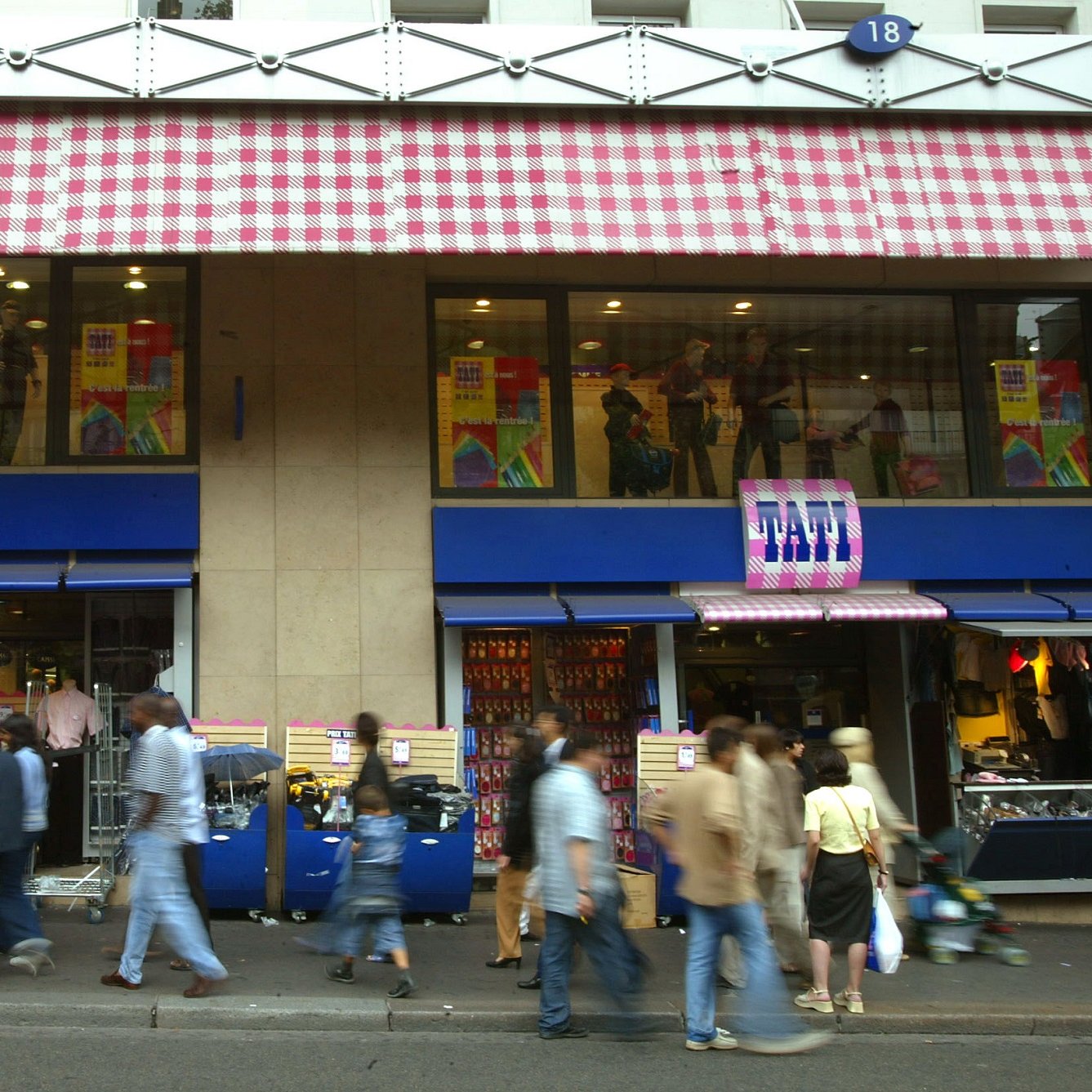 L'image montre une rue animée avec des piétons marchant devant un magasin. Le magasin est identifié par un enseigne "FATI" et présente des vitrines avec des mannequins habillés. Les façades supérieures du magasin ont des stores à carreaux. On peut voir une multitude de personnes se déplacer sur le trottoir, créant une atmosphère dynamique et vivante.