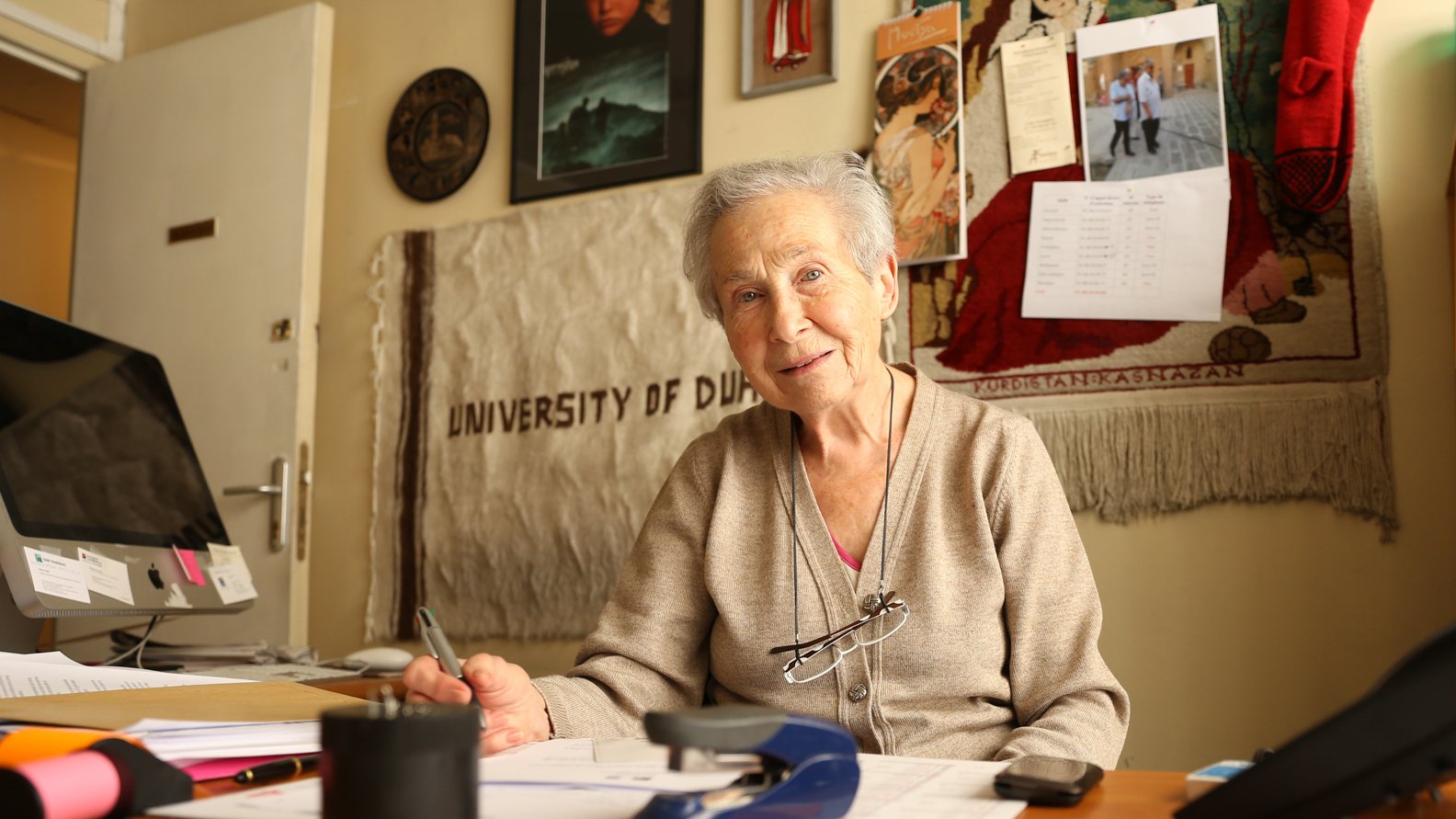 Femme âgée souriante, assise à un bureau avec des documents et des décorations murales.