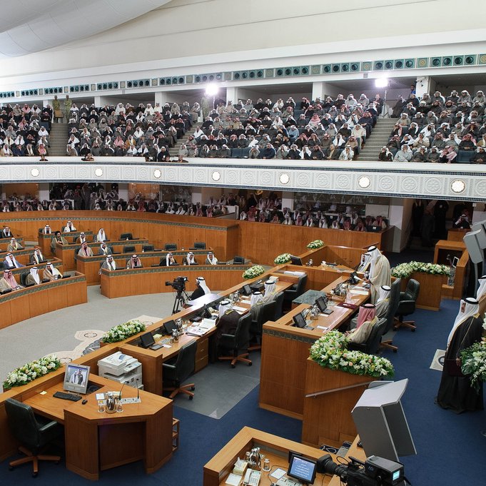 L'image montre une grande assemblée, probablement un parlement ou une salle de réunion officielle. On y voit des hommes en tenue traditionnelle, souvent vêtus de thobes et de keffiehs, assis à des bureaux en bois agencés en demi-cercle. La salle est bien aménagée, avec des décorations florales sur les tables et des écrans affichant des informations. Une grande audience est présente, ce qui suggère une session importante. L'ambiance semble solennelle et axée sur la discussion ou la prise de décision.