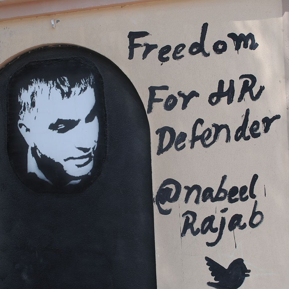 الصورة تظهر جدارية على حائط، تحتوي على صورة لشخص يبدو وكأنه إنسان. تحت الصورة، هناك نص مكتوب باللغة الإنجليزية يقول "حرية لمدافع حقوق الإنسان" مع ذكر اسم "@nabeel Rajab". الألوان المستخدمة تتضمن الأسود والأبيض، حيث تبرز الصورة بشكل يلفت الانتباه. تعبر الجدارية عن دعم لشخصية معروفة في مجال حقوق الإنسان.
