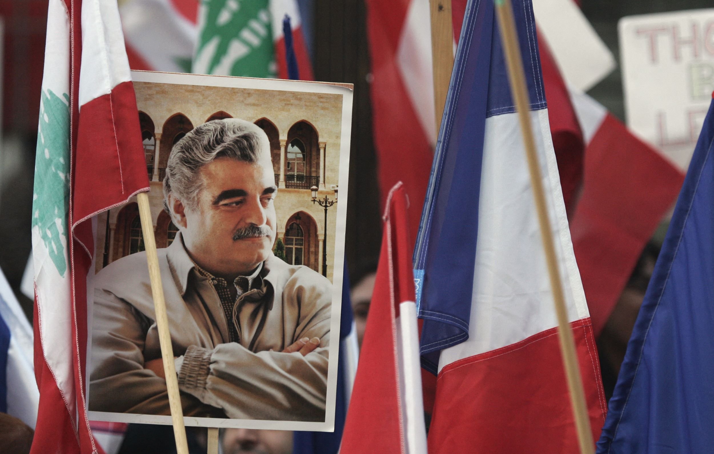 L'image montre une manifestation où plusieurs drapeaux, dont ceux du Liban et de la France, sont visibles. Au centre, il y a un portrait d'un homme, tenant une expression sérieuse, et entouré de personnes tenant des drapeaux. L'atmosphère semble chargée d'émotion et d'engagement.