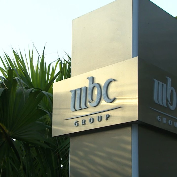L'image montre un panneau en métal portant le logo de "MBC Group". Le panneau est installé dans un environnement extérieur, entouré de végétation, notamment des palmiers. Le fond est clair, suggérant une ambiance ensoleillée.
