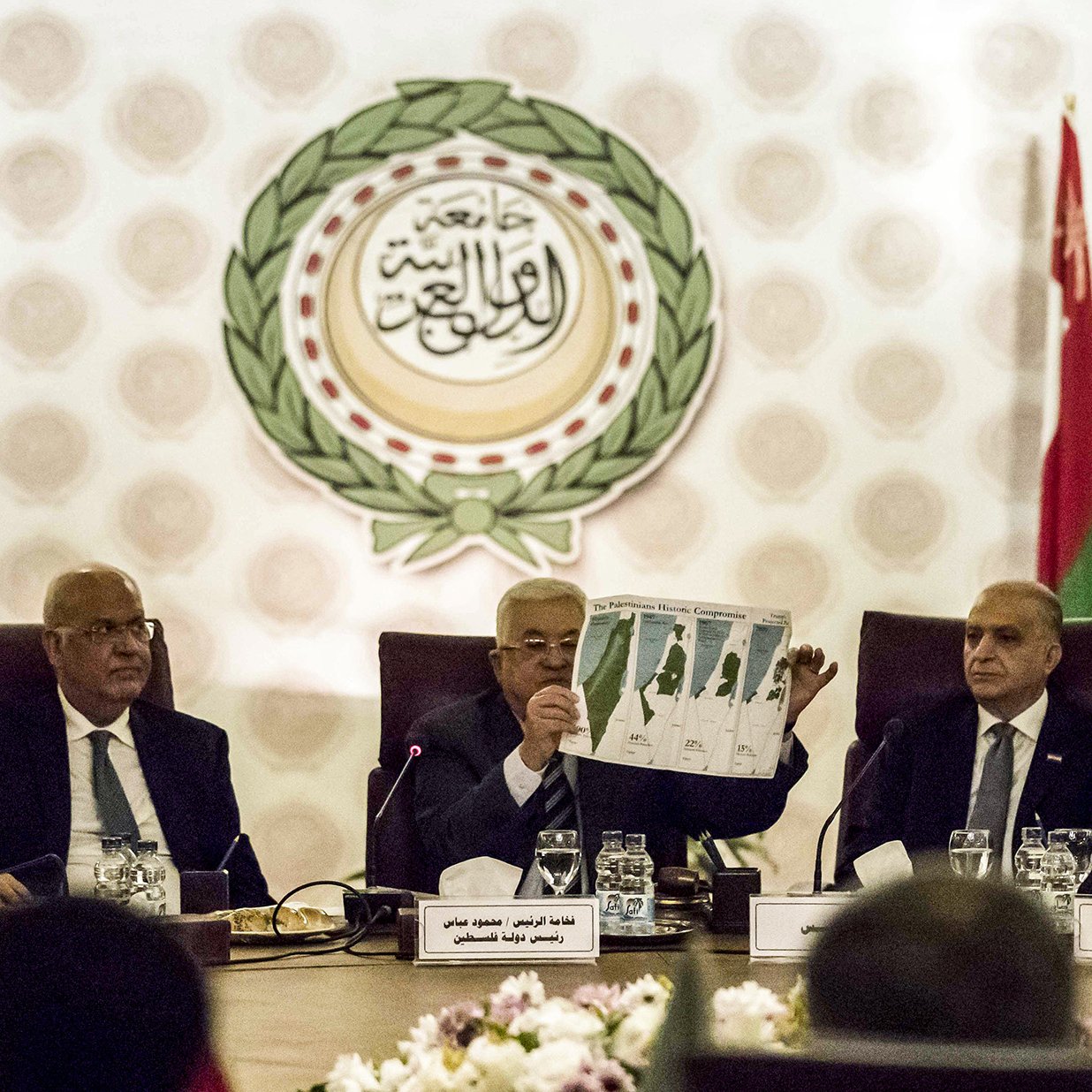 در این تصویر، گروهی از افراد در یک جلسه رسمی نشسته‌اند. یکی از افراد در حال نشان دادن یک نقشه یا نمودار به دیگران است. پشت آنها پرچم‌های کشورهای مختلف و نماد یا لوگوی اتحادیه عرب قابل مشاهده است. فضای جلسه حرفه‌ای و سازمان‌یافته به نظر می‌رسد. اعضای حاضر به دقت به موضوع مورد بحث گوش می‌دهند.