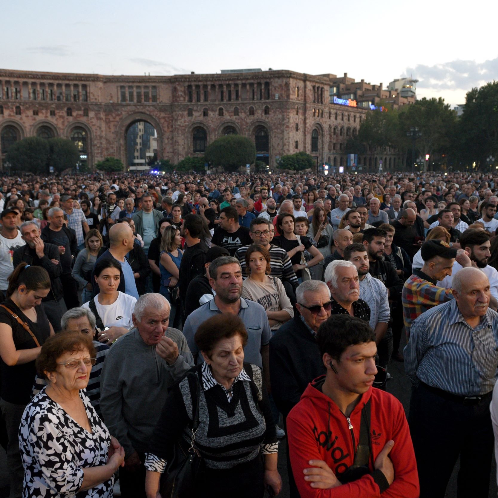 L'image montre une grande foule de personnes rassemblées dans un espace public, probablement pour un événement ou une manifestation. Les gens semblent concentrés et attentifs, certains regardant vers l'avant. L'arrière-plan présente des bâtiments, suggérant un milieu urbain, et le ciel est légèrement nuageux, indiquant que le jour commence à tomber. L'atmosphère semble à la fois solennelle et engagée.
