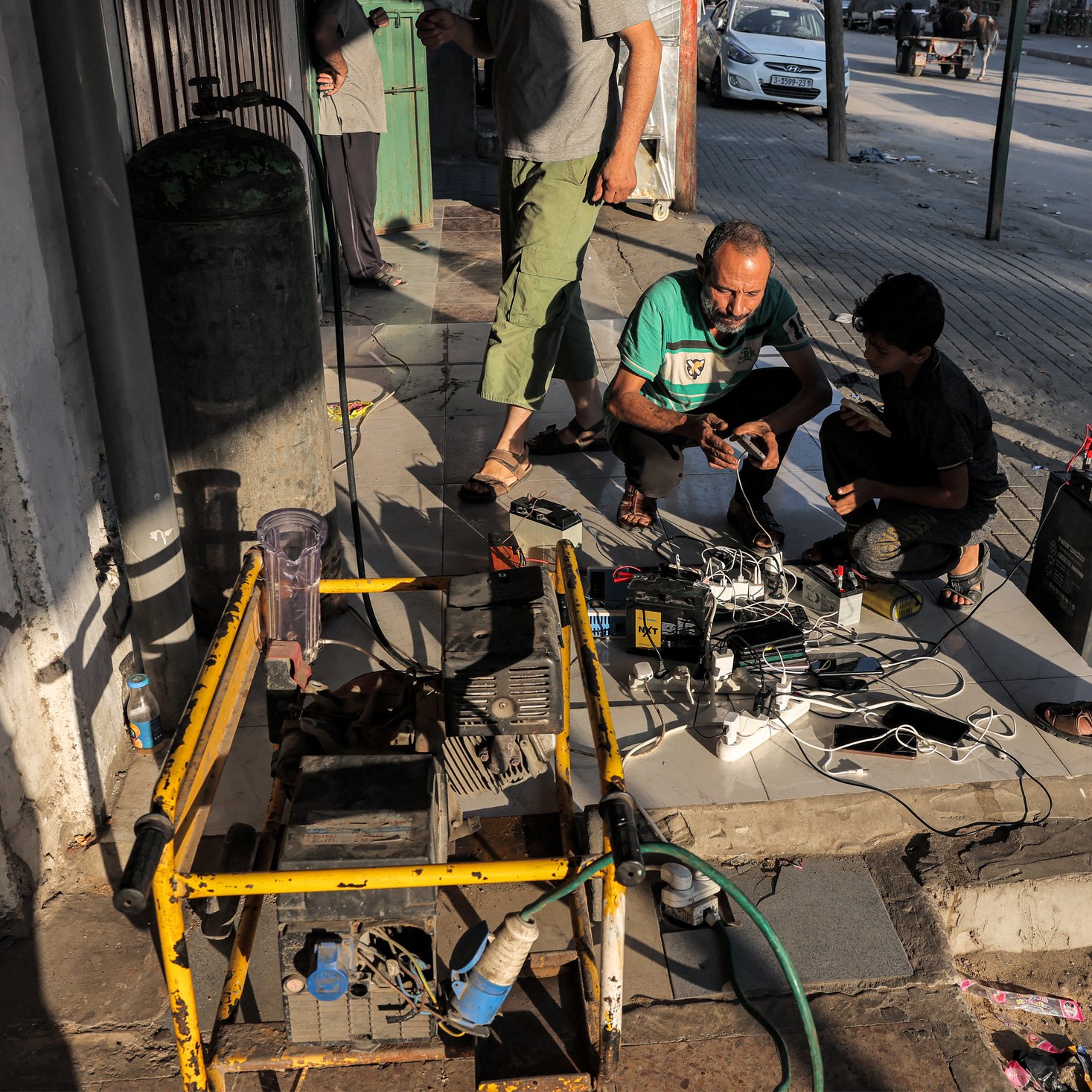 در تصویر، گروهی از افراد در حال کار بر روی یک دستگاه یا تجهیزات الکتریکی در خارج از یک ساختمان هستند. تعدادی سیم و کابل در اطراف آنها وجود دارد و به نظر می‌رسد که مشغول تعمیر یا بررسی یک دستگاه هستند. در پس‌زمینه، خیابانی دیده می‌شود و تعدادی وسیله نقلیه نیز در حال عبور هستند. جو تصویر به صورت غیررسمی و مشغول به کار به نظر می‌رسد.