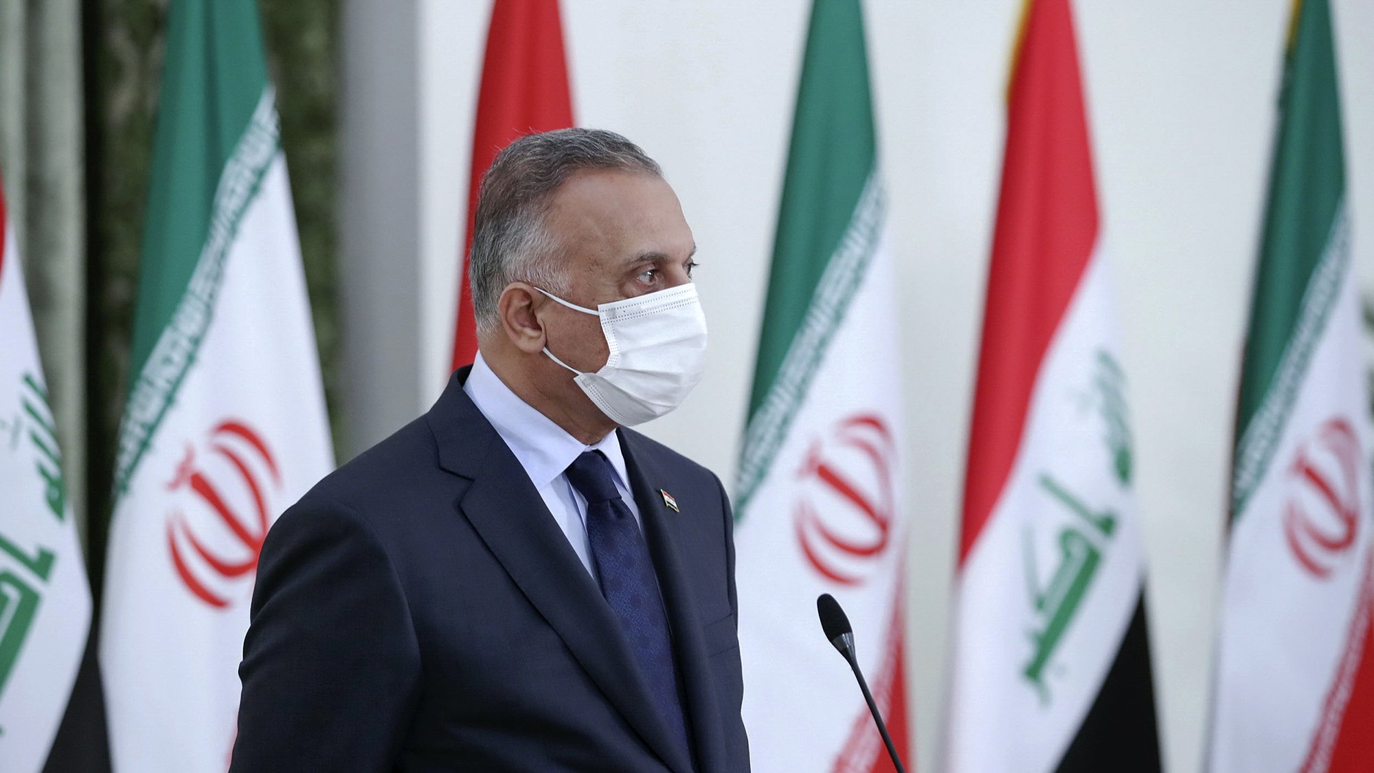 در این تصویر، مردی با روپوش رسمی و ماسک صورت دیده می‌شود. او در حال ایستاده و به یک سمت نگاه می‌کند. پشت او پرچم‌های ایران و عراق در ردیف قرار گرفته‌اند. فضا به نظر رسمی و جدی است، که نشان‌دهنده یک رویداد یا کنفرانس است.