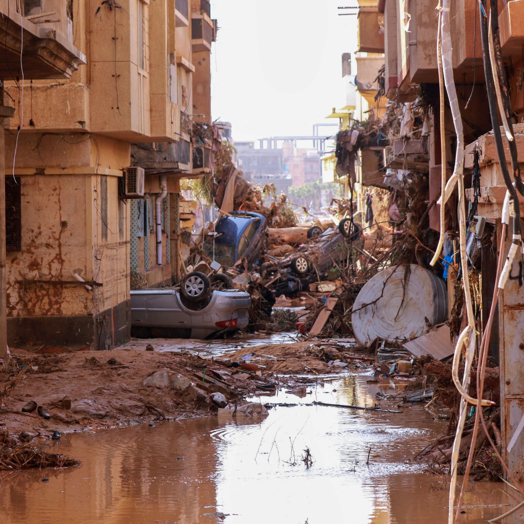 این تصویر یک خیابان خراب‌شده را نشان می‌دهد که آثار خسارت‌های ناشی از سیلاب یا طوفان بر روی ساختمان‌ها و وسایل نقلیه مشهود است. دیوارهای ساختمان‌ها آسیب دیده و برخی از آن‌ها بخشی از نمای خود را از دست داده‌اند. در وسط خیابان، آب گل‌آلودی جمع شده و در کنار آن، وسایل و اشیاء مختلفی مانند منازل و اتومبیل‌ها، به‌هم‌ریخته و به سمت خیابان آمده‌اند. این تصویر حال و هوای نگران‌کننده و ویرانگر ناشی از یک فاجعه طبیعی را به نمایش می‌گذارد.