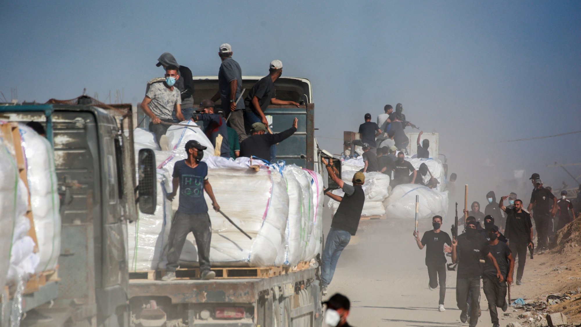 تصویر شامل کامیون‌هایی است که بار سنگینی حمل می‌کنند و عده‌ای در حال کار در یک محیط غبارآلود هستند.