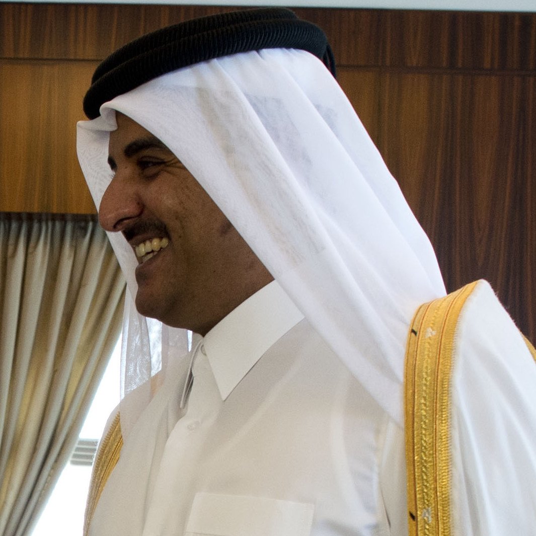 L'image montre un homme portant une tenue traditionnelle qatarienne. Il a un large sourire et semble être en mouvement, peut-être en train de parler ou d'interagir avec d'autres personnes. Sa tenue est composée d'une robe blanche, avec des ornements dorés, et il porte également un turban blanc avec un bandeau noir. L'arrière-plan montre des draperies et un environnement intérieur, indiquant un cadre formel ou officiel.
