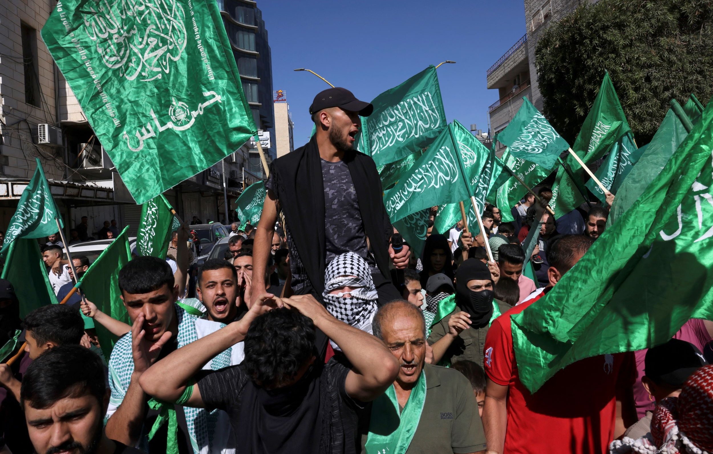 L'immagine mostra un gruppo di persone in una manifestazione. Molti partecipanti brandiscono bandiere verdi, che possono simboleggiare un movimento politico o religioso. Alcuni individui sembrano esprimere forti emozioni, con sorrisi e grida, mentre uno di loro è in piedi sopra la folla, probabilmente cercando di guidare o incoraggiare i manifestanti. Lo sfondo presenta edifici e un cielo sereno, suggerendo che l'evento si svolge all'aperto in una città.