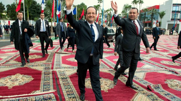 L'image montre deux hommes marchant sur un tapis richement décoré. L'un d'eux, vêtu d'un costume sombre et souriant, fait un geste de la main, tandis que l'autre, également en costume, marche à ses côtés. En arrière-plan, on peut voir des drapeaux de différentes nations, ce qui suggère un événement international ou officiel. L'atmosphère semble conviviale et solennelle à la fois.