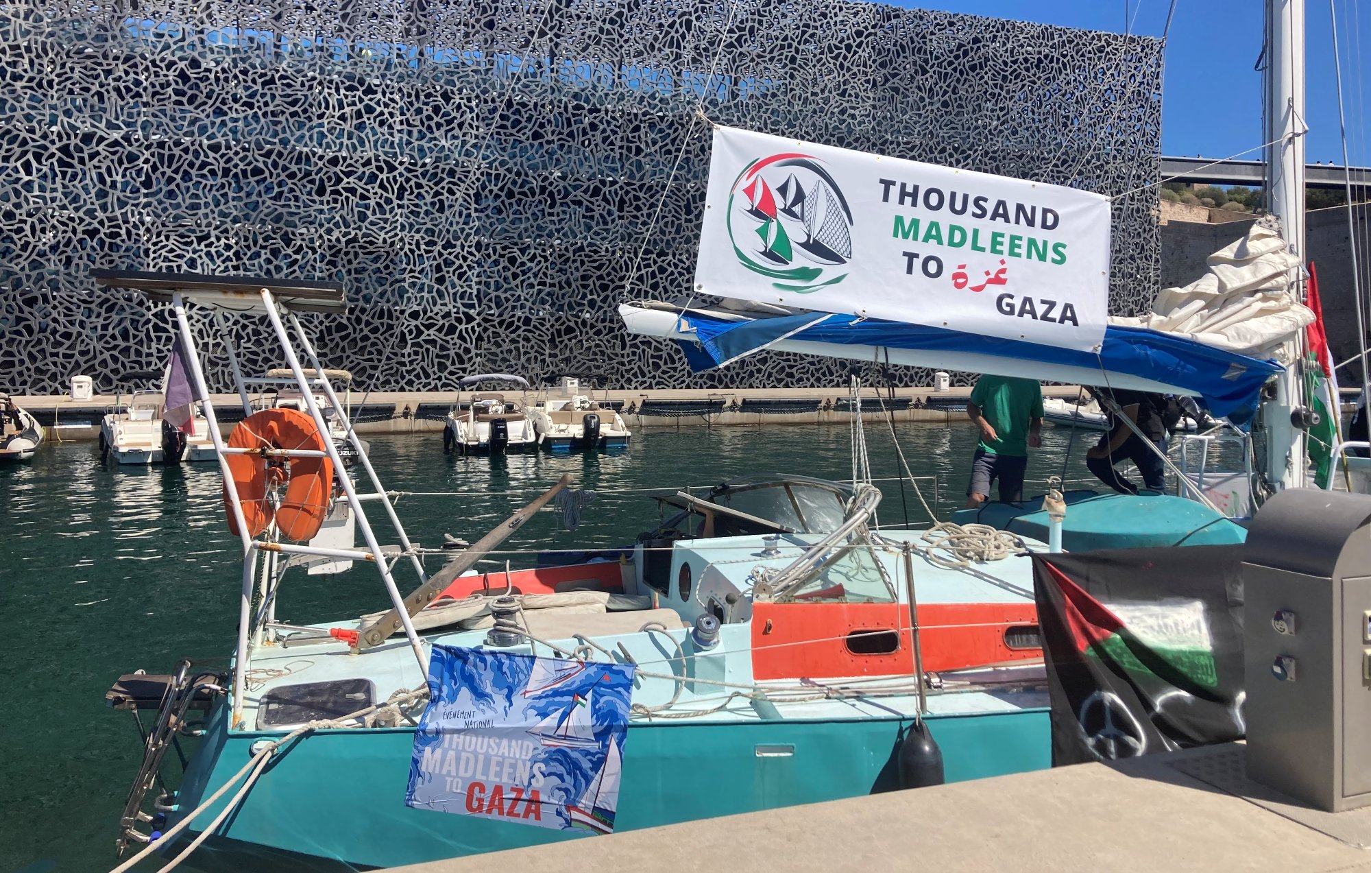 Un bateau avec une banderole "Thousand Madleen to Gaza" dans un port moderne.