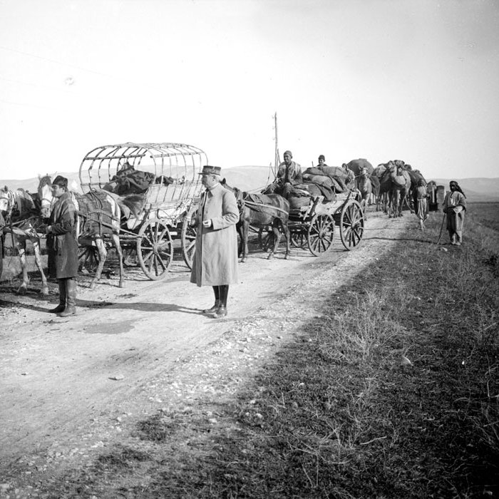 L'image montre une scène historique sur un chemin. On y voit plusieurs chariots tirés par des chevaux, ainsi que des personnes qui se déplacent. Certaines personnes sont debout près des chariots, tandis que d'autres semblent s'affairer autour des animaux. L'environnement est rural, avec des champs en arrière-plan et une atmosphère qui évoque des temps passés, probablement sur un chemin de terre. Les vêtements des personnes suggèrent une époque ancienne.