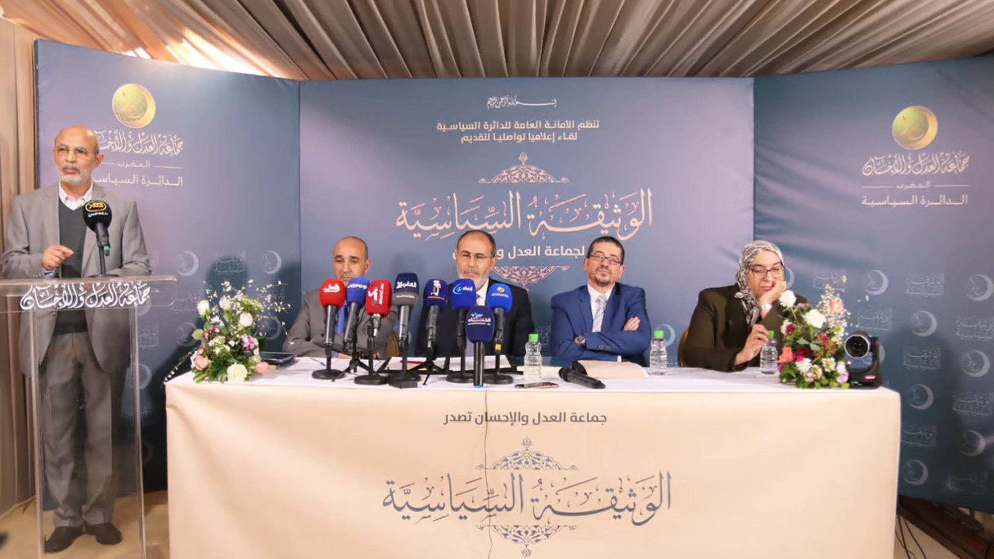 الصورة تظهر مؤتمرًا صحفيًا أو اجتماعًا رسميًا. يوجد على المنصة أربعة أشخاص يجلسون معًا، حيث يتحدث أحدهم ويبدو أنه يتحدث في ميكروفون. هناك لافتة خلفهم تحمل عنوان الاجتماع، ومجموعة من الكاميرات والشاشات تسجل أو تنقل الحدث. الطاولة مزينة بالزهور، والإضاءة هادئة. الخلفية تبدو أنيقة وتعكس الجدية الرسمية للاجتماع.