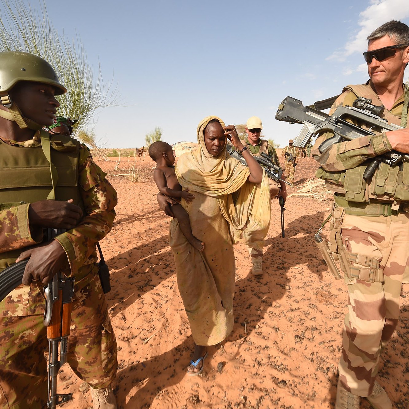 L'image illustre une scène de terrain militaire dans un environnement désertique. On peut voir deux soldats armés, l'un en uniforme de camouflage avec un casque, et l'autre portant un uniforme militaire beige. Ils escortent une femme vêtue d'une robe traditionnelle, qui porte un enfant dans ses bras. L'arrière-plan montre un paysage désertique avec quelques arbres, suggérant une zone aride. Cette image évoque des thèmes de sécurité, de protection et d'interaction humanitaire.