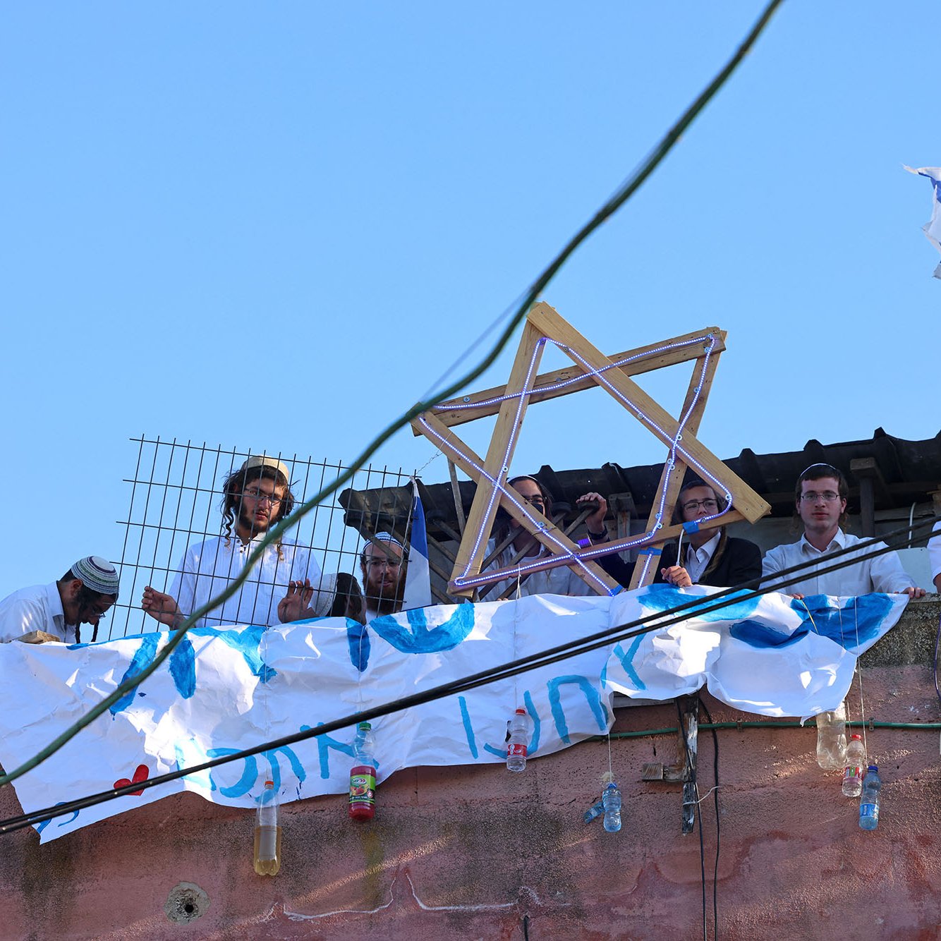 L'image montre un groupe de personnes rassemblées sur un toit. Elles portent des vêtements traditionnels, et certaines tiennent un symbole en forme d'étoile de David. En arrière-plan, il y a un drapeau israélien flottant. On peut également voir une banderole accrochée, suggérant un message ou une revendication. Le ciel est dégagé, et il y a une ambiance de rassemblement communautaire.