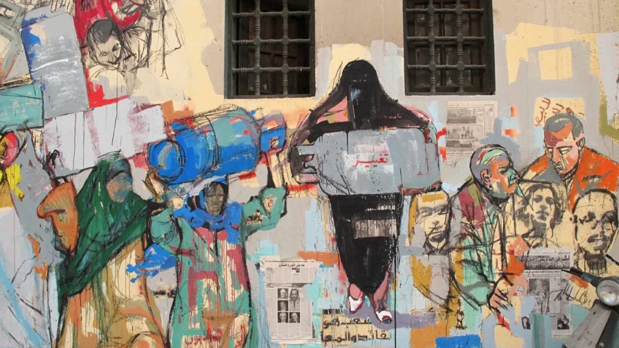 L'image montre une fresque murale colorée qui illustre diverses figures humaines engagées dans des activités sociales et politiques. Les personnages semblent porter des objets, portant des expressions variées, ce qui évoque des thèmes de lutte et de solidarité. Le mur est également décoré de collage de papiers et de textes, ajoutant une dimension visuelle riche et narrative à l'œuvre. Les couleurs utilisées sont vibrantes et contrastées, ce qui attire l'attention sur les détails et les émotions des personnages représentés.