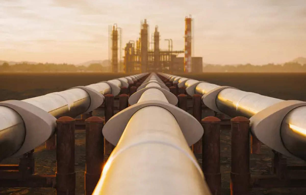 L'image montre une vue d'un long pipeline qui s'étend vers une installation industrielle en arrière-plan. Les tuyaux sont disposés sur des supports et semblent converger vers l'usine, éclairée par une lumière dorée, probablement au lever ou au coucher du soleil. L'environnement autour est vaste et dégagé, avec des montagnes lointaines à l'horizon, créant une ambiance industrielle mais aussi naturelle.