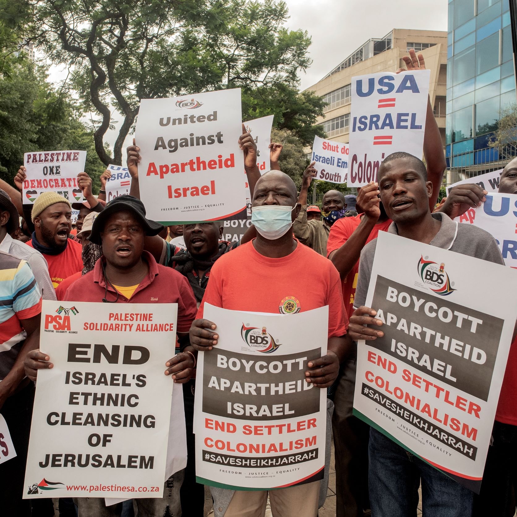 L'image montre une foule de manifestants tenant des pancartes et exprimant leur opposition à la politique israélienne, en particulier en ce qui concerne le traitement des Palestiniens. Les slogans sur les pancartes incluent des appels à mettre fin à l'apartheid et au colonialisme en Israël, ainsi qu'à boycotter certaines actions liées à ce pays. Les manifestants semblent très engagés dans leur cause, et l'atmosphère est dynamique et déterminée.