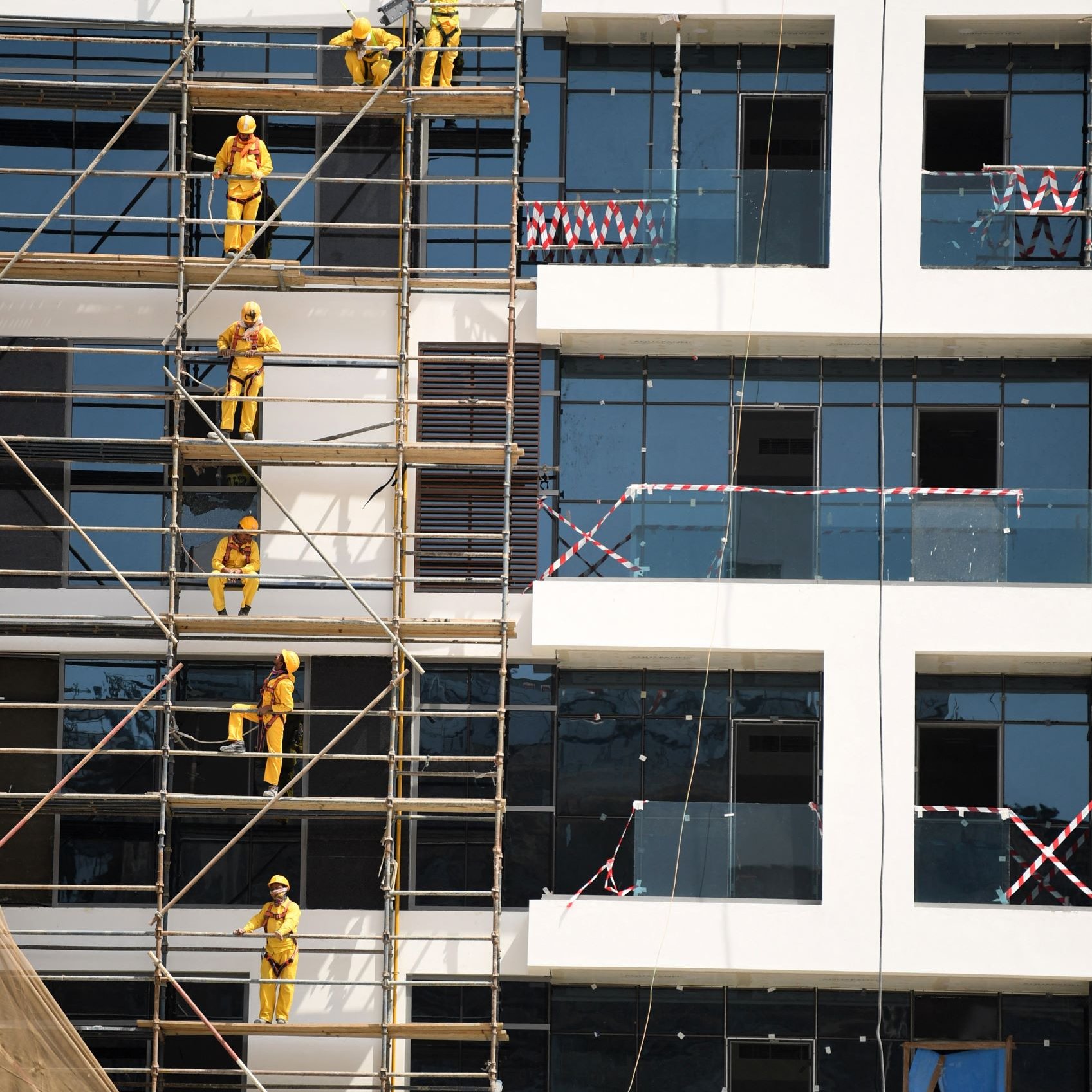L'image montre un bâtiment en construction. On peut voir des échafaudages le long de la façade, sur lesquels plusieurs ouvriers vêtus de combinaisons jaunes et de casques de sécurité sont en train de travailler. Certains utilisent des outils pour peindre ou poser des éléments sur les fenêtres, tandis que d'autres montent et descendent sur les échafaudages. Le bâtiment a des fenêtres en verre, et des bandes de sécurité sont visibles autour de certaines zones. L'ensemble de la scène reflète un environnement de travail actif et organisé.