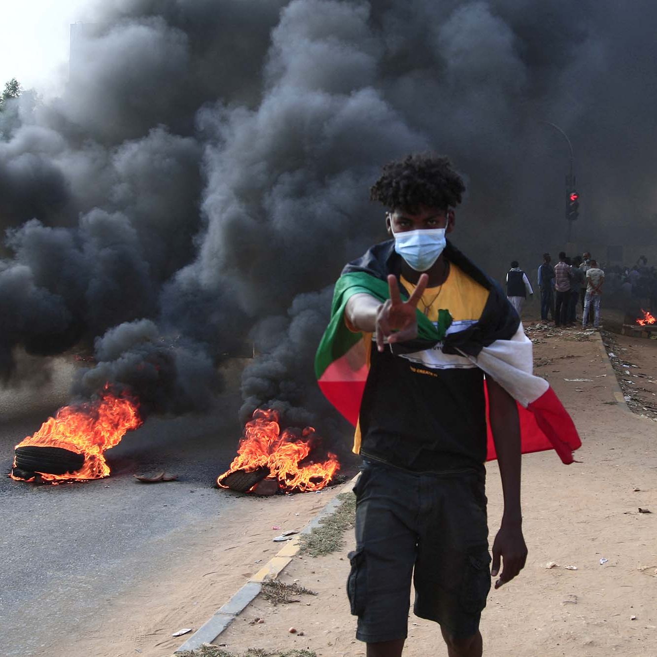 L'image montre un jeune homme marchant sur une route pendant une manifestation. Il porte un masque et fait un signe de paix avec sa main. En arrière-plan, on peut voir des pneus enflammés et une épaisse colonne de fumée noire s'élevant dans l'air. La scène dégage une impression de tension et de protestation, et il y a un groupe de personnes visibles derrière lui.