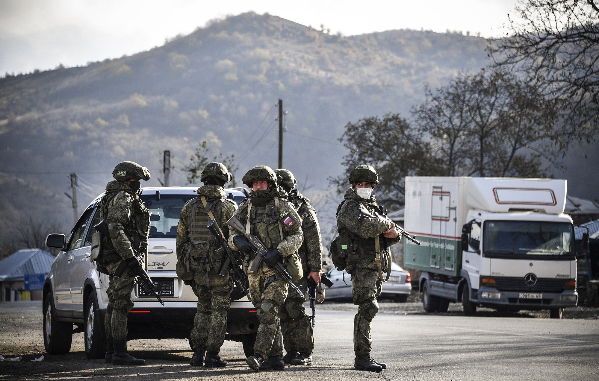 L'image montre un groupe de soldats armés en uniforme, se tenant près d'un véhicule. Ils semblent être en position de vigilance, avec des montagnes en arrière-plan. L'atmosphère est sérieuse, suggérant une opération militaire ou une situation de sécurité. Des camions peuvent également être aperçus à proximité, ajoutant un contexte logistique à la scène.