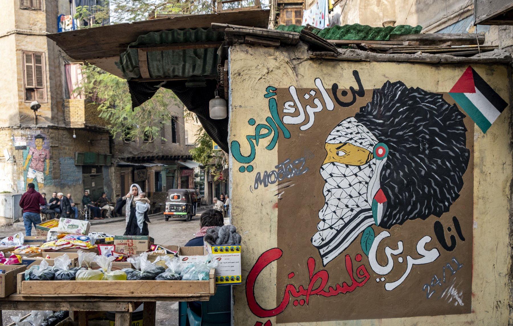 L'image montre un mur peint avec une fresque représentant une figure symbolique, probablement inspirée par la culture palestinienne. Le personnage porte un keffieh et a une chevelure élaborée. À côté, il y a des inscriptions en arabe qui expriment des sentiments de résistance et d'identité palestinienne. En avant-plan, il y a un marché avec des étals de fruits et légumes, et une rue animée où l'on peut apercevoir des passants et des véhicules. L'ambiance reflète un mélange de culture, d'art et de vie urbaine.
